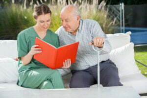 Senior man reading book with female caretaker on couch at nursing home
